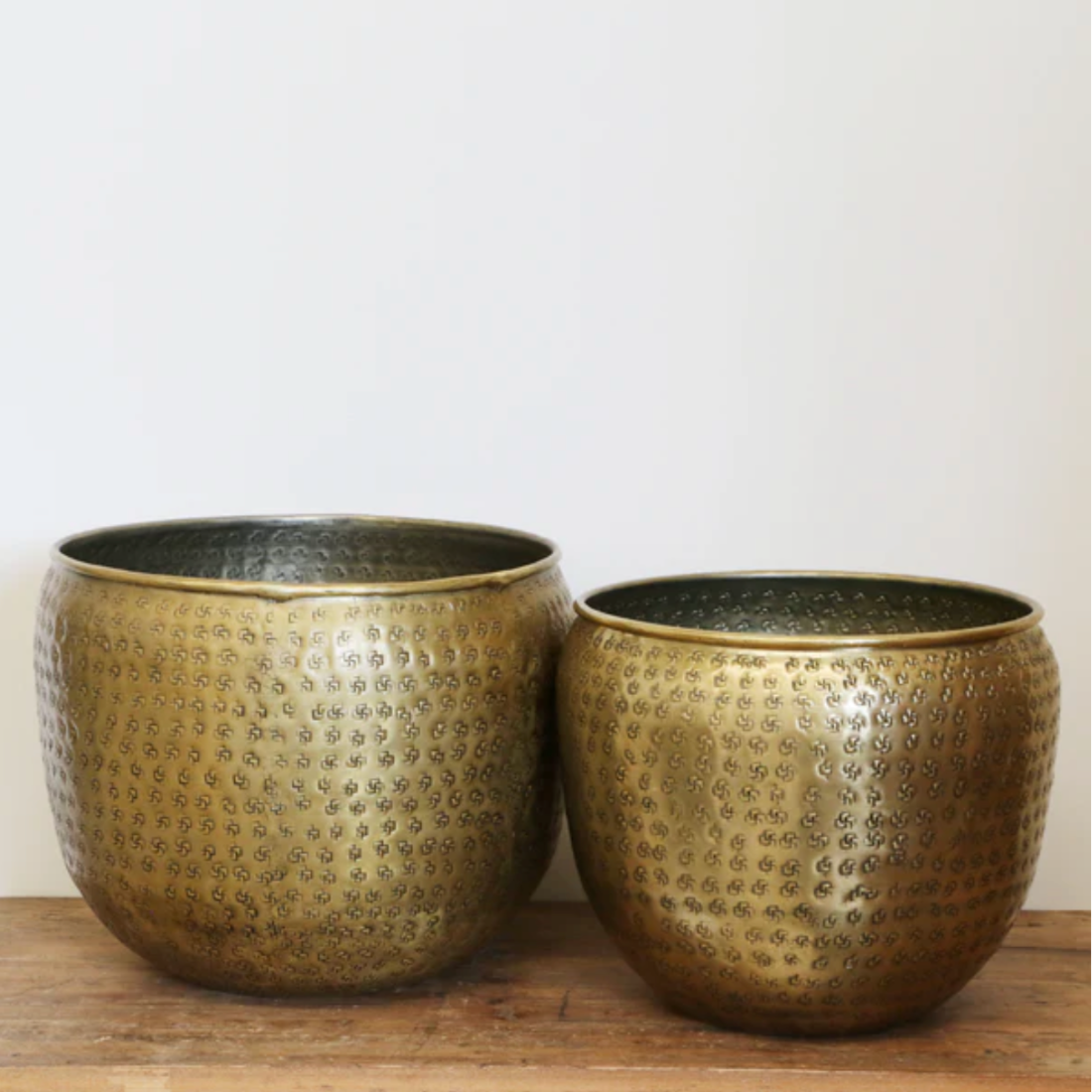 Two antique brass textured planters on a wooden surface with a plain background