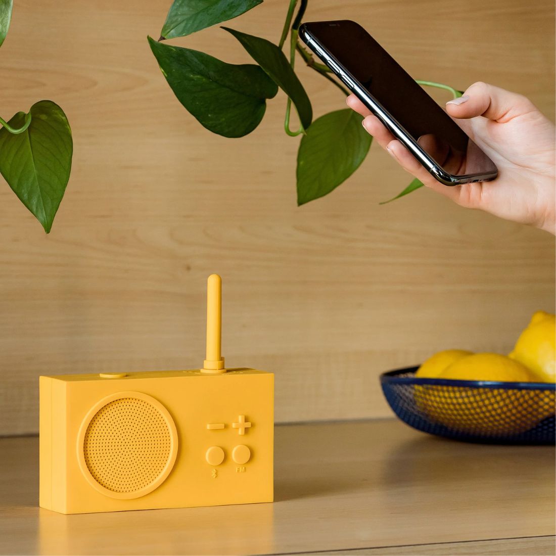 Yellow portable radio on a wooden surface with a hand holding a smartphone and a bowl of lemons in the background.
