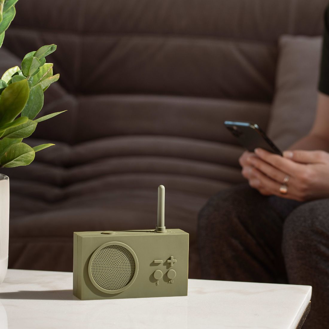 Khaki green retro-style portable radio on a table with a person using a phone to play music on the radio in the background