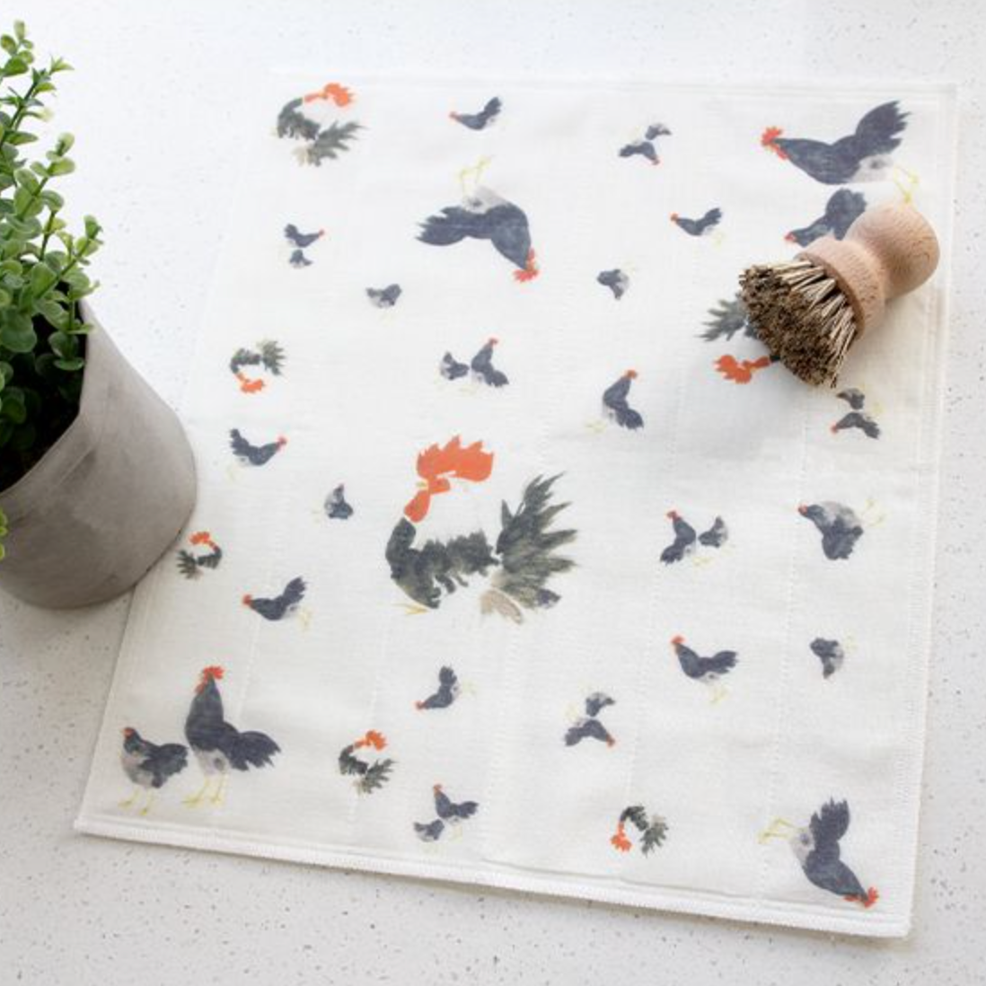 Dishcloth with chicken pattern on a white surface next to a plant and a plant pot.