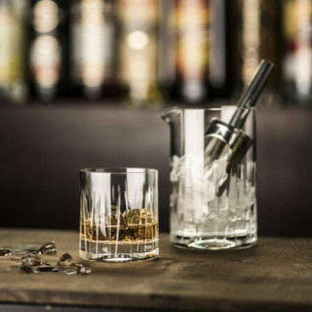Bar setup with a glass of whiskey and a cocktail shaker on a wooden surface, with blurred bottles in the background.